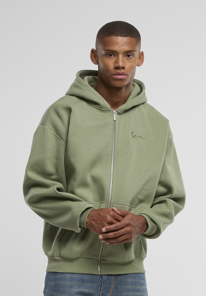 Jeune homme portant un sweat à capuche zippé vert clair, debout les mains jointes, devant un fond gris clair uni.