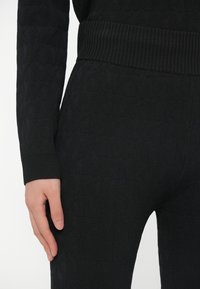 Pulover negru tricotat, cu model texturat, manșete și talie reiate. Prim-plan cu braț și detaliu al materialului cu un luciu subtil.