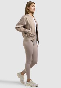 Kvinde iført beige bomberjakke, hvid crop top, beige leggings og hvide sneakers, stående foran en lysegrå baggrund.