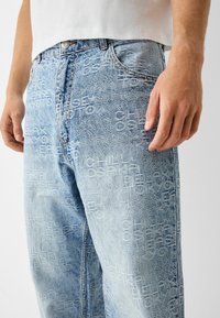 Ljusblå denimjeans med präglade textmönster. Designen inkluderar en vanlig midja och fem fickor, med en avslappnad passform.