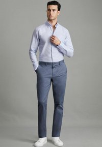 Lichtblauw overhemd met lange mouwen en een kraag, gecombineerd met marineblauwe broek en witte sneakers, tegen een neutrale achtergrond.