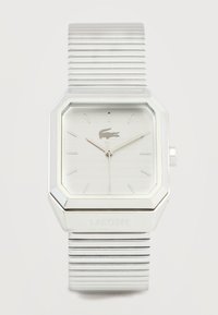 Ασημένιο τετράγωνο ρολόι χειρός Lacoste με κυματοειδές μεταλλικό λουράκι, μίνιμαλ δείκτες καντράν και λογότυπο κροκοδείλου στη θέση 12 η ώρα.