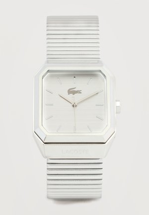 Strieborné štvorcové hodinky Lacoste s rebrovaným kovovým remienkom, s minimálnymi značkami na ciferníku a logom krokodíla na pozícii 12 hodín.