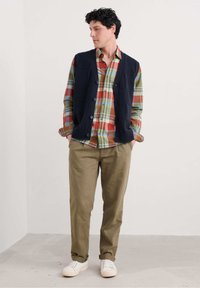 Marine gebreide vest over een multicolored geruit shirt; tan broek en witte sneakers. Gladde textuur, knoopdetails, casual pasvorm.