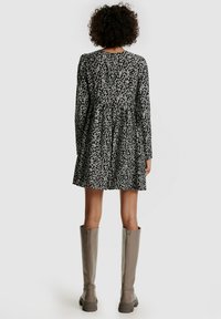 Schwarzes Blumenkleid mit langen Ärmeln und einem ausgestellten Rock, aus strukturiertem Stoff, mit weißen Blumenmustern. Kombiniert mit knielangen grauen Stiefeln.