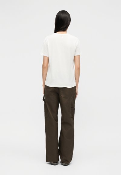Γυναίκα με μακριά σκούρα μαλλιά που φοράει μια απλή λευκή κοντομάνικη μπλούζα και φαρδιά σκούρα καφέ παντελόνια cargo, όρθια με την πλάτη στραμμένη.