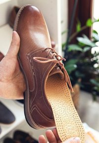 Brown leather shoe with perforated upper, lace-up design, tan insole featuring holes, and a rubber sole. Hand holding the shoe.