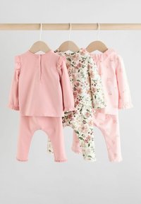 Drei Sets Kinderbekleidung hängen an einer Holzstange. Das erste ist einfarbig rosa mit Rüschen, das zweite hat ein Blumenmuster und das dritte ist hellrosa mit zarten Blumen.
