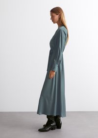 Lang, teal kjole med belte i midjen, med lange ermer og en glatt tekstur, paret med svarte hælboots.