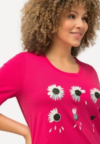 Mujer con cabello rizado que lleva una camisa de color rosa brillante con gráficos de margaritas blancas con centros negros y un pequeño detalle de corazón negro.