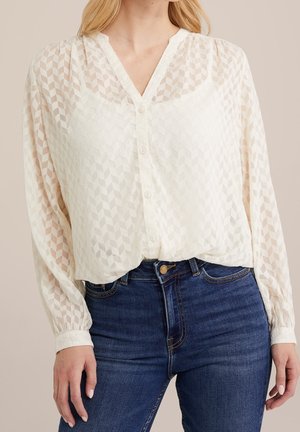 Vrouw draagt een witte blouse met knopen en een geometrisch patroon en blauwe spijkerbroek, staand tegen een effen achtergrond.