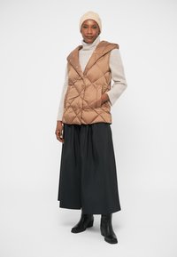 Veste matelassée marron avec capuche, motif en losanges, associée à un pull léger et une longue jupe noire. Des bottines noires complètent la tenue.