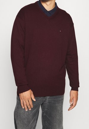 Mand iført en mørkerød V-hals sweater over en navy skjorte med krave og falmede grå jeans, stående med hænderne afslappet ned langs siderne.