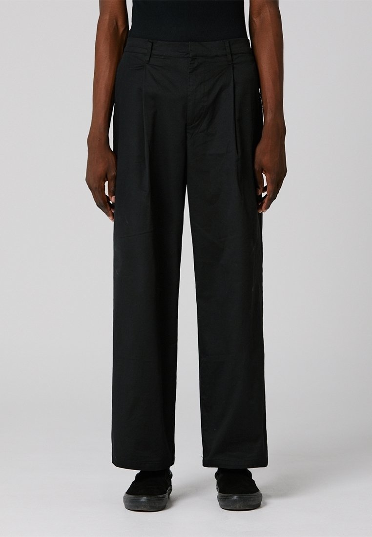 Pantalons larges noirs en tissu lisse. Comprend des plis à l'avant, une fermeture éclair et des poches latérales. Taille texturée avec passants de ceinture.