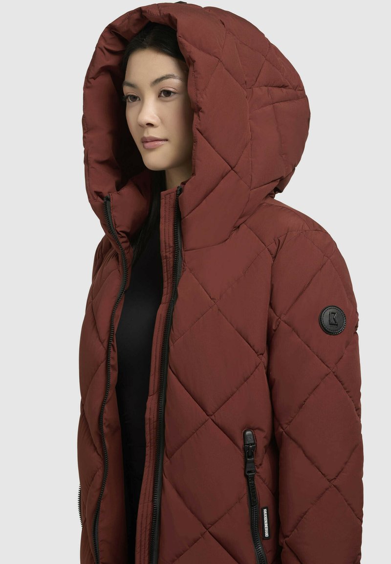 Gesteppt, ziegelroter Pufferjacke mit großer Kapuze. Verfügt über schwarze Reißverschlüsse und ein kreisförmiges Logo-Patch am linken Ärmel.