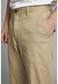 Beige bavlněné kalhoty mají plochý přední díl, boční kapsy a zapínání na knoflík. Tkanina má hladkou texturu s mírným leskem.