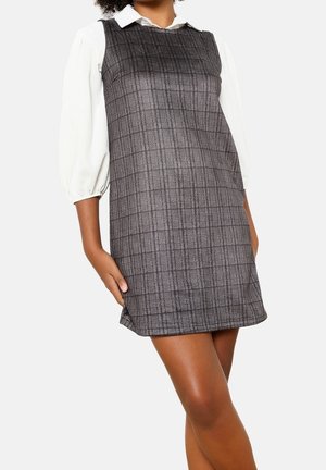 Frau trägt ein graues, kariertes ärmelloses Kleid über einer weißen Bluse mit Puffärmeln, steht mit einer Hand auf dem Oberschenkel vor einfarbigem Hintergrund.