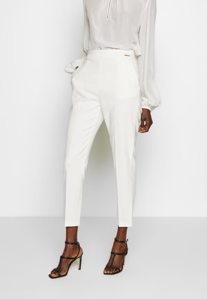 Femme portant un pantalon blanc sur mesure longueur cheville, un chemisier blanc transparent à manches longues, et des sandales à talons hauts noires à brides, debout.