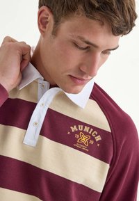 Mørkerød og creme-stribet poloshirt med hvid krave. Har et logo med teksten "MUNICH" og året 1939 samt dekorative knapper på kraven.