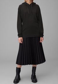 Czarna bluza z kapturem wykonana z miękkiego materiału, w połączeniu z plisowaną czarną midi spódnicą oraz czarnymi botkami na kostkę z grubą podeszwą.