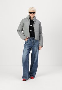 Giacca imbottita grigia con colletto alto, maglione nero con cuori bianchi, jeans blu a gamba larga e scarpe rosse. Tessuto morbido e vestibilità comoda.