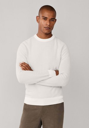 Suéter blanco de punto con mangas largas, escote redondo y puños acanalados, combinado con pantalones oscuros de textura marrón. Diseño minimalista.