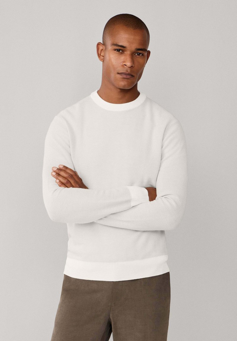 Maglione bianco a maglia con maniche lunghe, scollatura rotonda e polsini a costine, abbinato a pantaloni strutturati marrone scuro. Design minimalista.