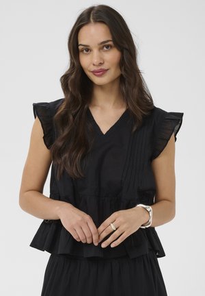 Femme aux longs cheveux bruns ondulés portant un haut noir sans manches avec des épaules à volants et un bracelet argenté, sur un fond uni.