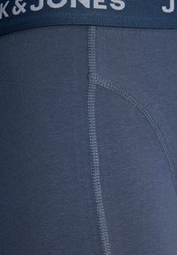 Moške temno modre boksarice z mehkimi elastičnimi pasom, ki vsebujejo belo besedilo logotipa. Gladka tkanina z vidnimi šivi.