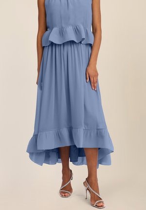 Frau in einem ärmellosen blauen Midikleid mit Rüschen und silbernen High-Heel-Sandalen, stehend vor einem beigen Hintergrund.