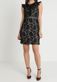 Robe en dentelle noire avec motifs floraux, manches courtes transparentes et taille ajustée. Finition avec un ourlet texturé, associée à des talons bordeaux.