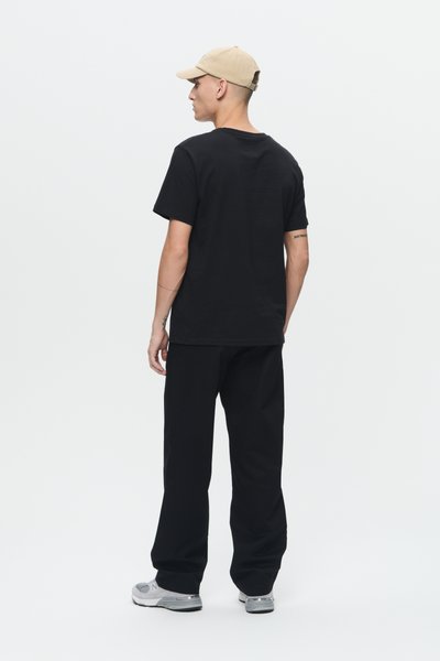 Camiseta de algodón negra de manga corta, combinada con pantalones negros holgados y zapatillas grises. Se lleva una gorra marrón hacia atrás.