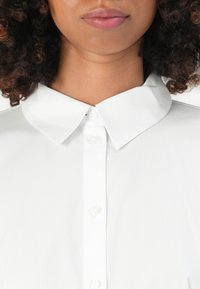 Primo piano della parte inferiore del viso e del collo di una persona con capelli ricci, che indossa una camicia bianca button-up ben stirata con colletto a punta.