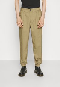 Beige, tettsydde bukser med folder og elastiske manchetter. Brukt med svarte støvler, med knappelukking og beltehemper. Glatt tekstur.