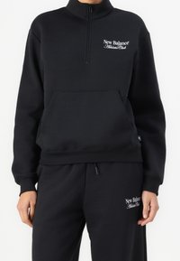 Zwarte fleece pullover met een ritskraag, voorzak en wit logo "New Balance Athletic Club" op de borst. Gladde textuur.