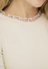 Krämfärgad ribbad topp med en delikat spetsvolang längs halsringningen, som visar en mjuk textur med en subtil och lätt design.