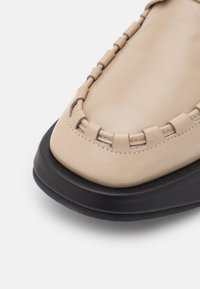 Mocassin en cuir beige avec des détails cousus à la main et une épaisse semelle en caoutchouc noire, présenté en gros plan sur un fond blanc.
