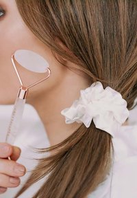 Biały jadeitowy roller do twarzy z metalową ramą i różowymi akcentami w kolorze złota. Gładka tekstura; trzymany przez osobę z białą gumką we włosach.