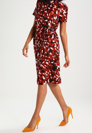 Mujer que lleva un vestido hasta la rodilla con un estampado abstracto en rojo, negro y blanco y un lazo en la cintura, combinado con zapatos de tacón alto de color naranja brillante.