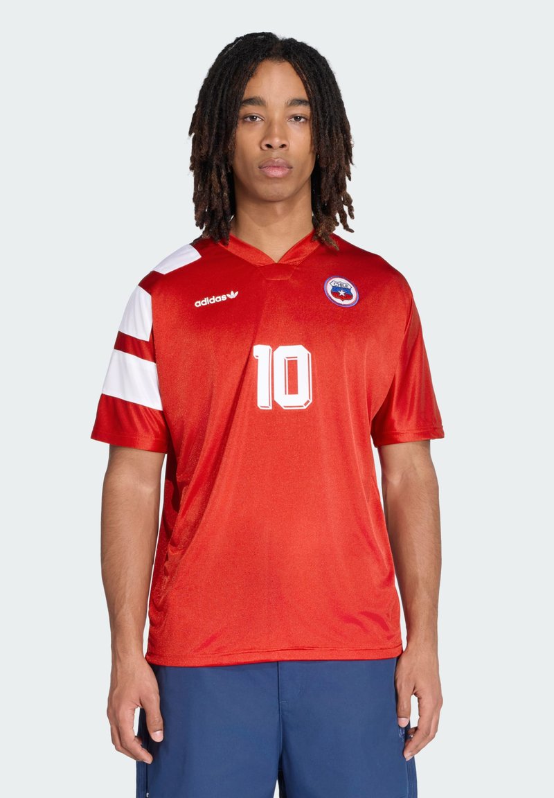 Jeune homme aux dreadlocks portant un maillot de football rouge avec le numéro 10, l'écusson du Chili, et un short bleu, debout devant un fond uni.