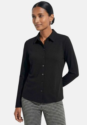 Femme portant une chemise noire à manches longues boutonnée et un pantalon pied-de-poule noir et blanc, debout avec une main derrière le dos.