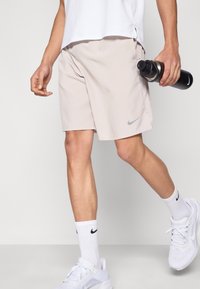 Vaaleanbeiget Nike-housut harmaalla logolla, yhdistettynä valkoisiin urheilusukkiin ja kenkiin. Oikeassa kädessä pidetään mustaa vesipulloa.