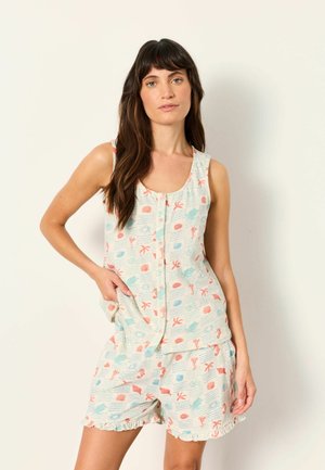 Frau trägt ärmelloses, geknöpftes Oberteil und passende Shorts mit pastellfarbenen Meerestier- und Korallenmotiven, steht vor einfachem Hintergrund.