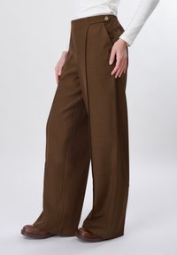 Femme portant un pantalon marron taille haute à jambes larges avec détail de boutons, associé à des chaussures marron et un haut blanc à manches longues.