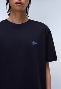 Tricou din bumbac de culoare bleumarin, cu un mic emblemă a unei steaguri albastre pe partea stângă a pieptului. Gât rotund, mâneci scurte și textură netedă.