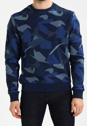 Mann trägt ein marineblaues Sweatshirt mit abstrakten geometrischen Mustern in verschiedenen Blautönen und weißen Punkten.