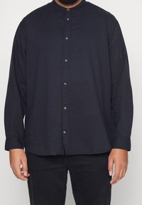 Uomo che indossa una camicia nera a maniche lunghe con colletto alla mandarina e pantaloni neri, in piedi contro uno sfondo semplice.