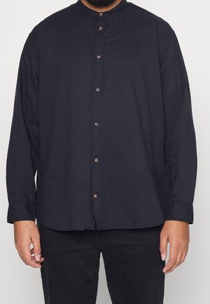 Man in een zwart overhemd met lange mouwen en een opstaande kraag, gecombineerd met zwarte broek, staand tegen een effen achtergrond.