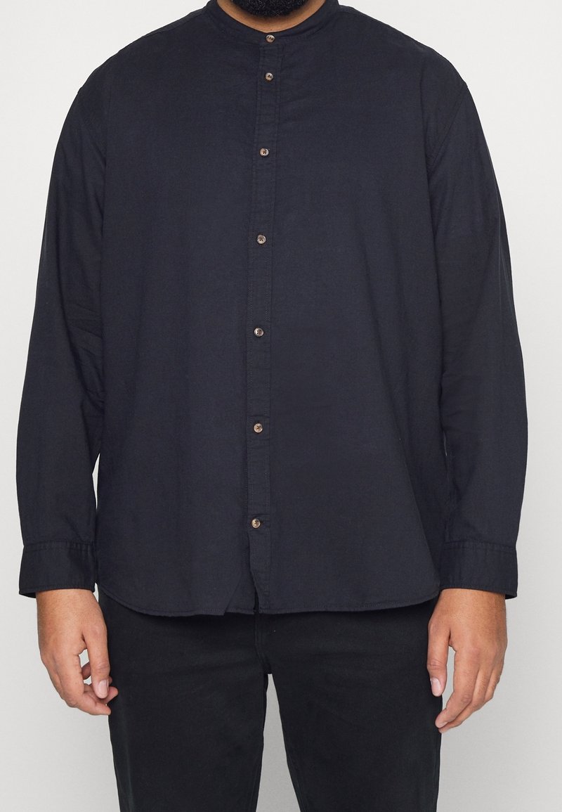 Uomo che indossa una camicia nera a maniche lunghe con colletto alla mandarina e pantaloni neri, in piedi contro uno sfondo semplice.