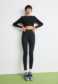 Kvinna i svart långärmad crop top, högmidjade svarta leggings, vita strumpor och silverfärgade Nike-sneakers som står mot vit vägg på grön golv.